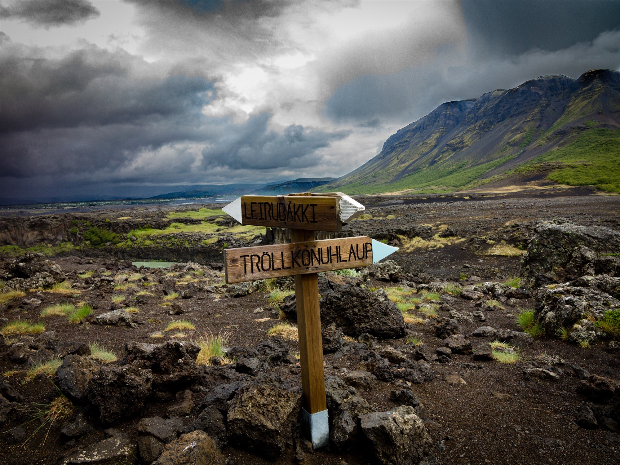 A brown wooden sign with Icelandic place names in front of a dramatic rocky landscape.