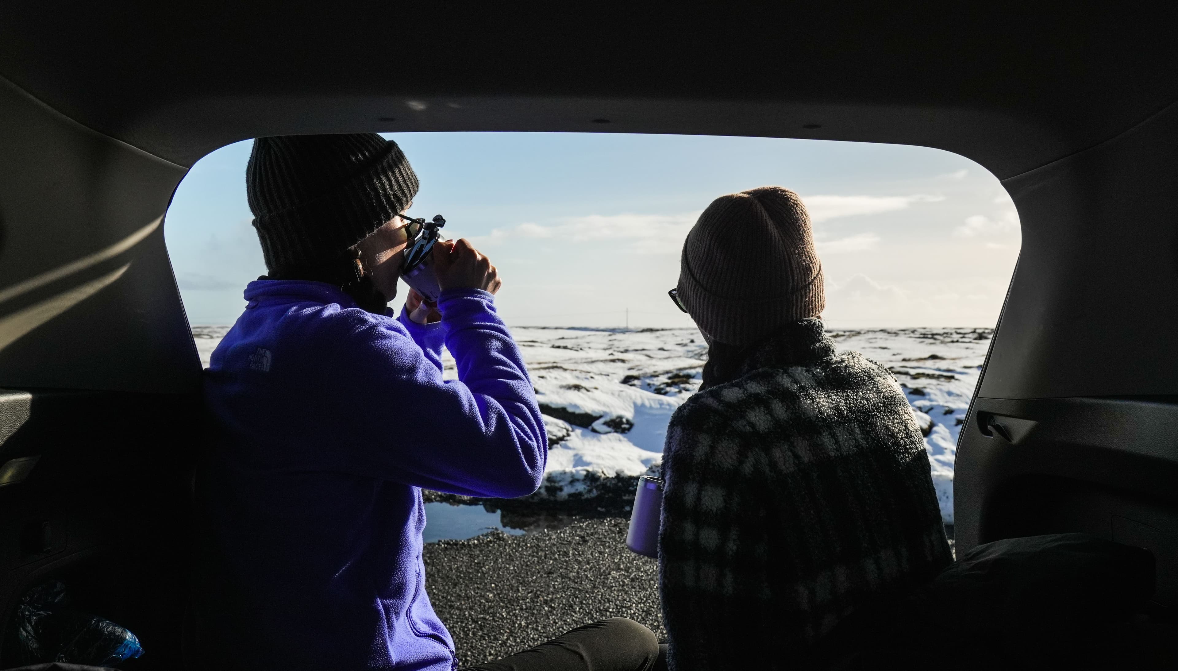 Image of two travelers in warm clothes drinking coffee in the back of a car.