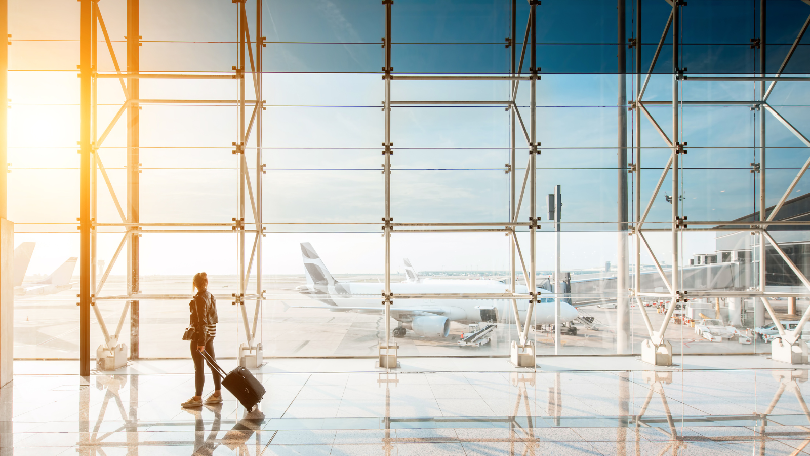 A woman pulling a trolley bag at an airport with floor-to-ceiling windows showing an aeroplane against gentle sunshine and blue skies in the background.