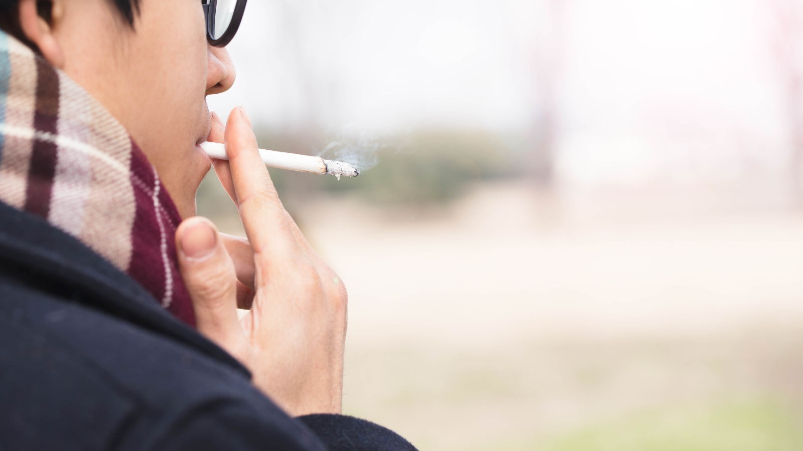 Man in glasses smoking outside in a coat and scarf.