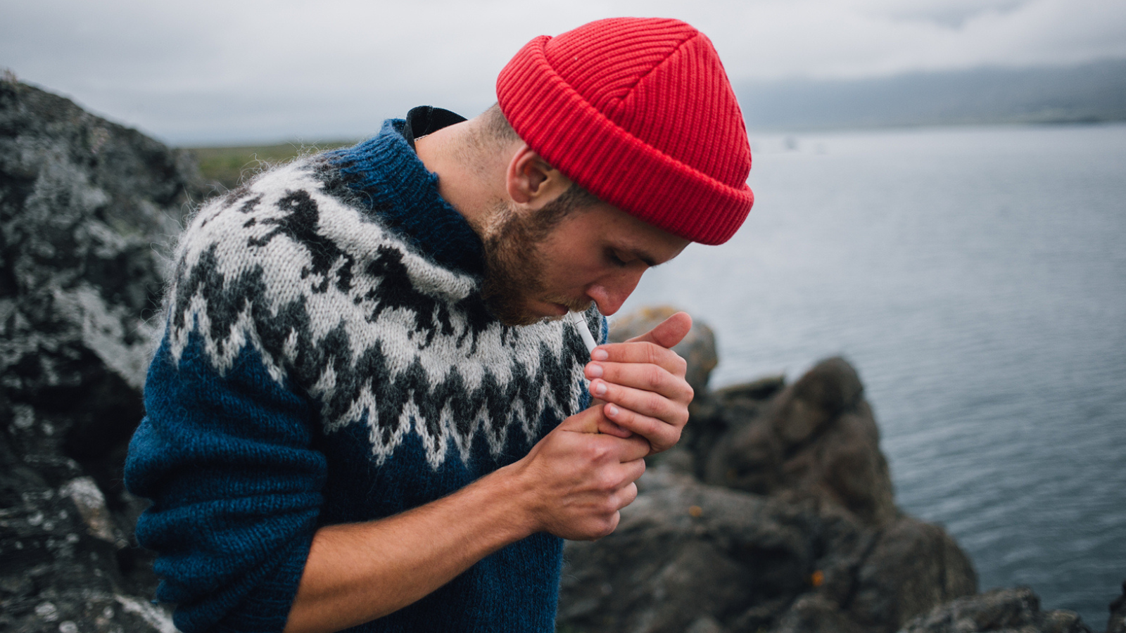 Man in an Icelandic sweater lighting a cigarette in front of a body of water.